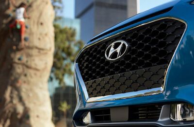 A close-up of the front of a blue Hyundai Kona, with the Hyundai logo prominently displayed on the grille. A rock climbing wall and cityscape are blurred in the background.