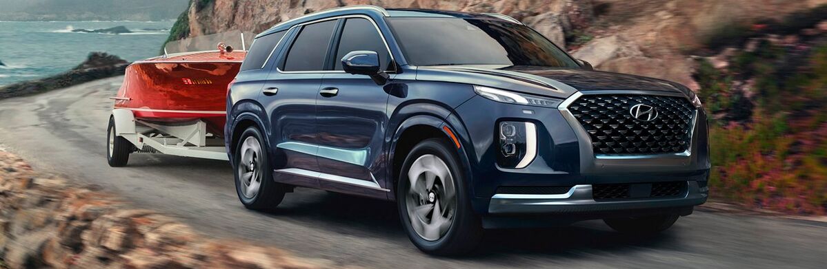 Dark blue Hyundai Palisade SUV towing a bright red trailer on a winding road with ocean views.