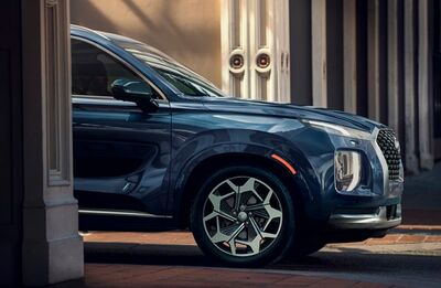 Close-up of the front fender and wheel of a blue Hyundai Palisade, with the front door slightly ajar, revealing a glimpse of the interior and a blurred urban backdrop.