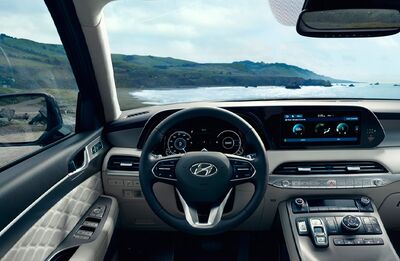 Interior view of a Hyundai Palisade dashboard, showing the steering wheel, instrument panel, and infotainment screen, with a scenic coastal view through the windshield.