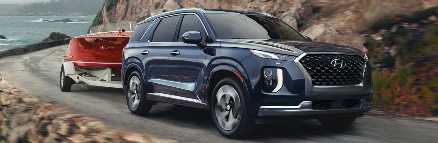 A blue Hyundai Palisade SUV driving along a winding coastal road, towing a vintage wooden boat behind it.