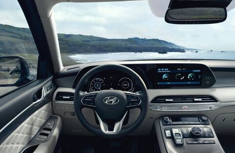 The interior of a Hyundai Palisade, showing the driver's seat, steering wheel, and dashboard with a large touchscreen display. A scenic coastal view is visible through the windshield.