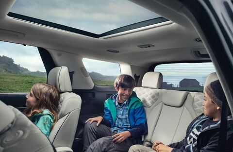 The interior of a Hyundai Palisade showing three children smiling and looking at each other while sitting in the back seats. A large panoramic sunroof is open above them.