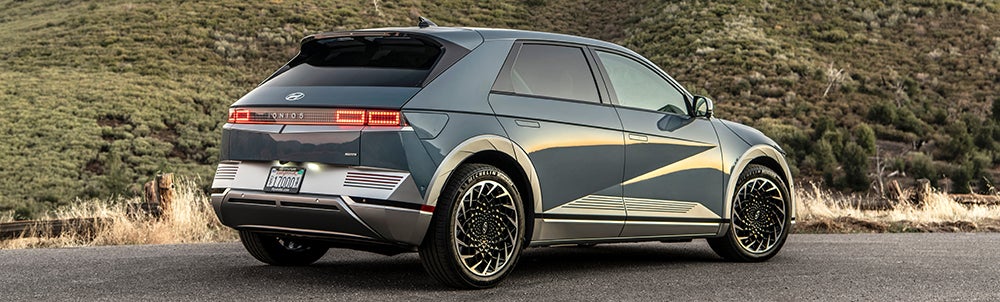 A rear three-quarter view of a blue Hyundai Ioniq 5 parked on a scenic road with hills and mountains in the background. The image highlights the car's distinctive pixelated taillights and futuristic design.
