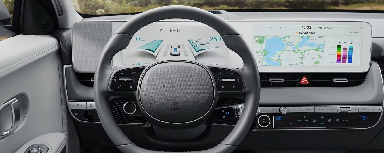 The interior of a Hyundai Ioniq 5, showcasing the driver's perspective. The image highlights the steering wheel, dashboard with a large touchscreen display, and a digital instrument cluster displaying vehicle information.