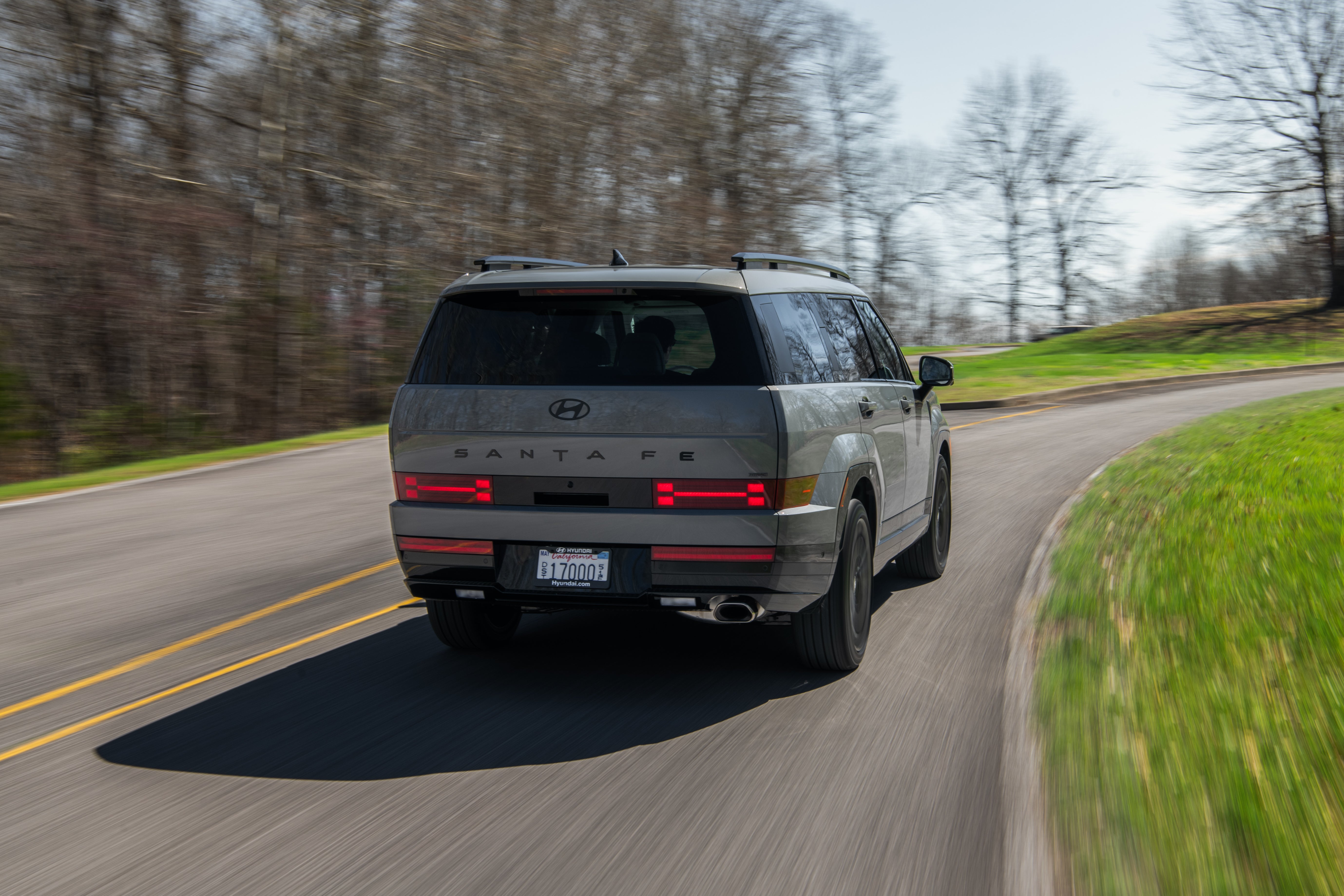 A rear view of a silver Hyundai Santa Fe driving on a winding road through a forest.
