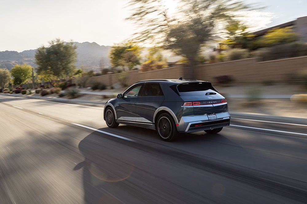 A dark gray Hyundai IONIQ 5 EV in motion on a sunny, winding road with a mountainous backdrop.