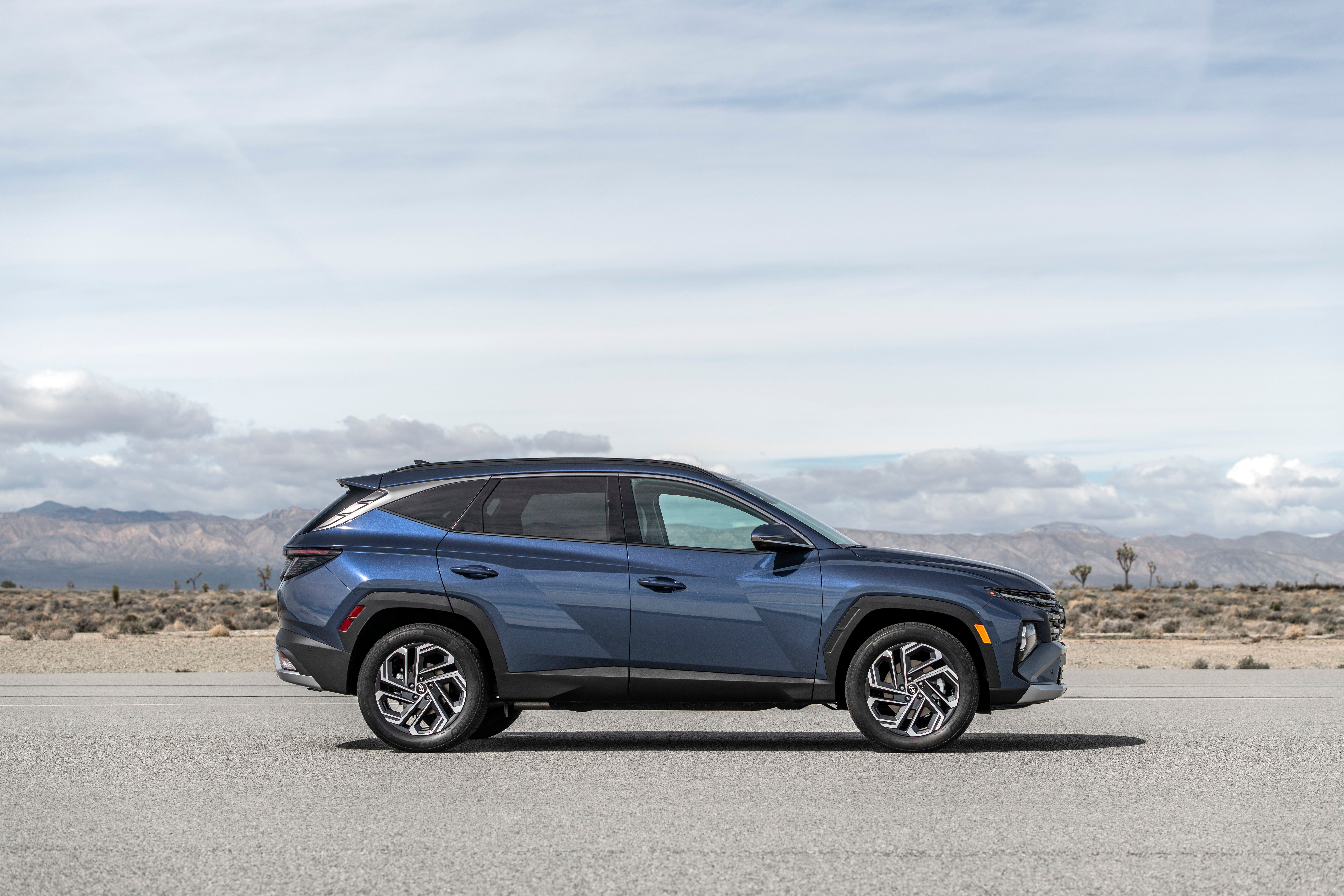 A side view of a blue 2026 Hyundai Tucson driving on a faded two lane asphalt road in a desert environment.