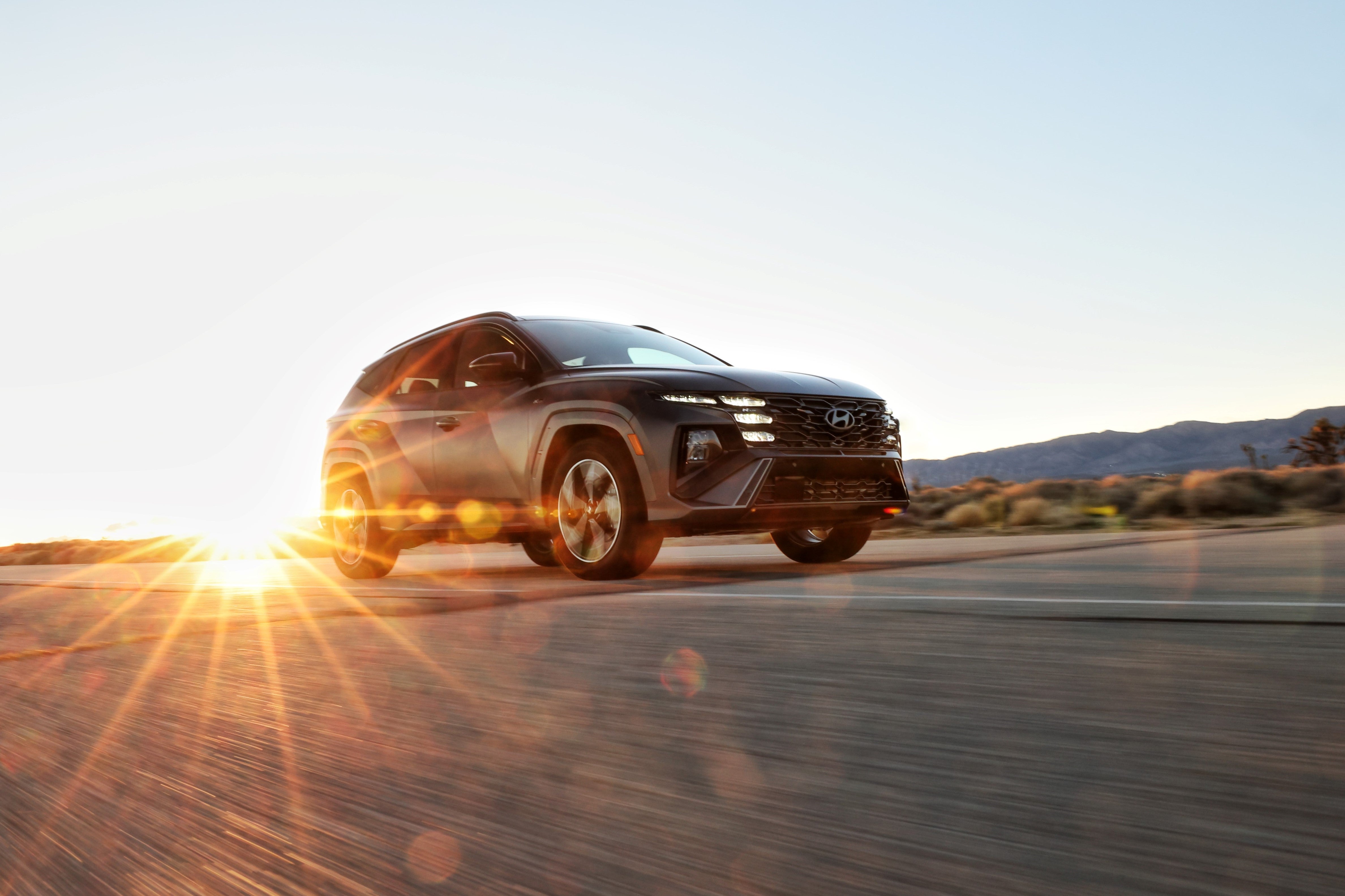 A grey new Hyundai Tucson Hybrid in Phoenix drives up a sloped desert road to the right with sunset light glaring from behind the vehicle.