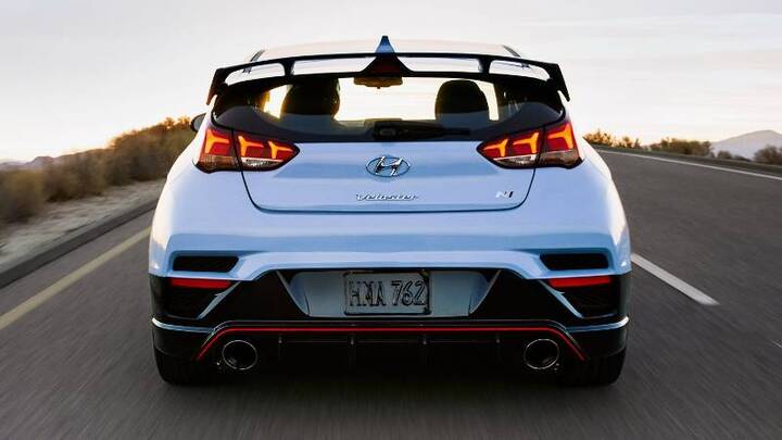 A rear view of a white Hyundai Veloster N driving on a highway at sunset. The car's distinctive rear spoiler, taillights, and dual exhaust tips are visible.