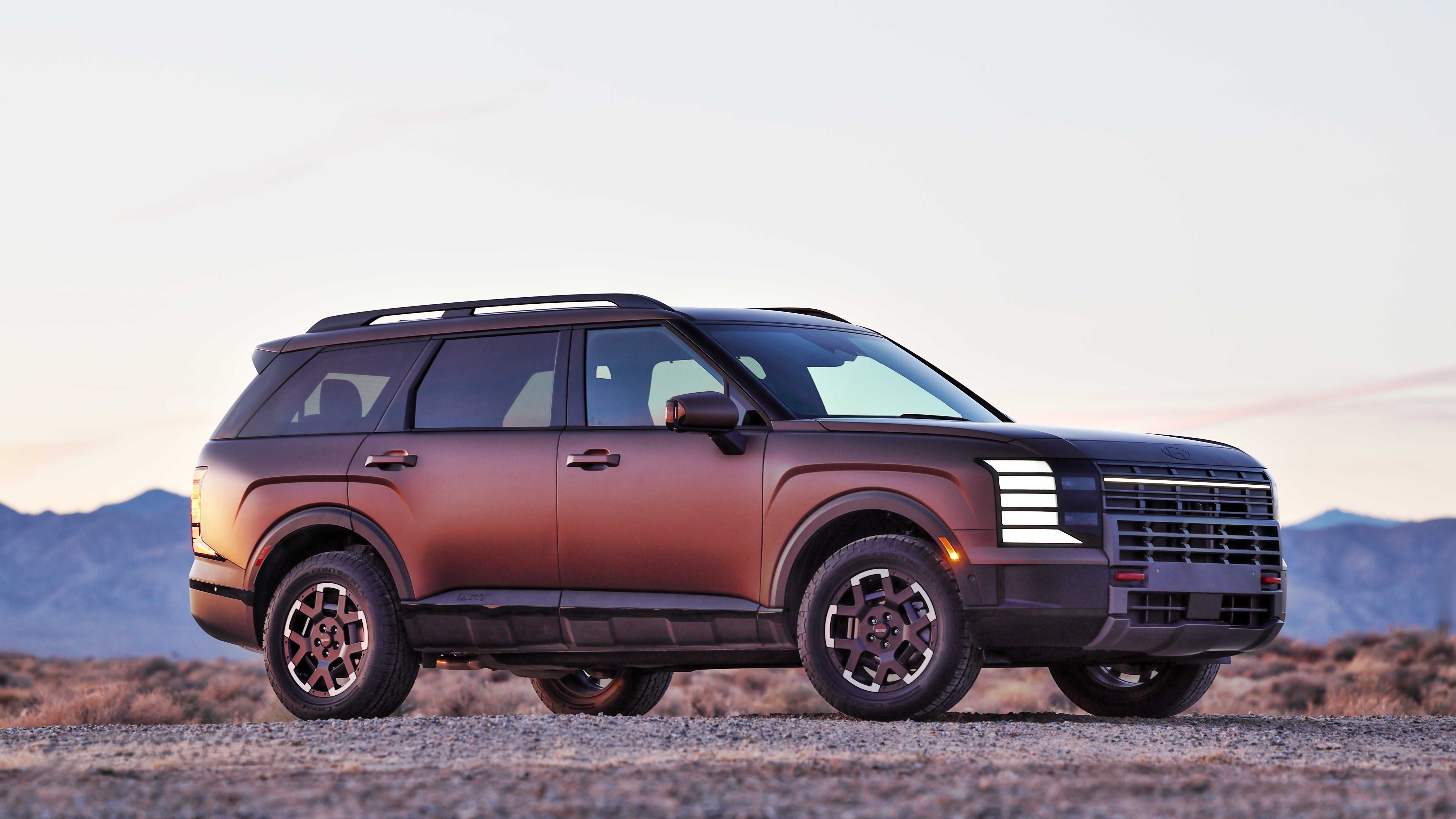 A bronze 2026 Hyundai Palisade in Scottsdale is parked facing the viewer to the right on a gravel surface with desert mountains in the background at sunset.