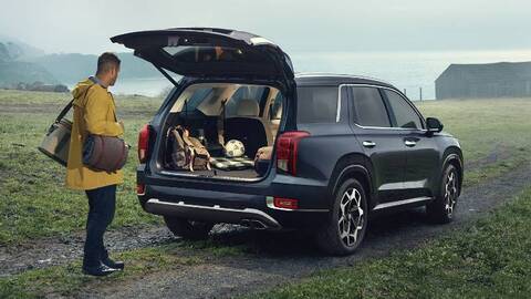 A comparison image of the Hyundai Palisade (left) and the Jeep Grand Cherokee L (right). The Palisade is parked in front of a bakery in a city setting, while the Grand Cherokee L is parked in a forest.