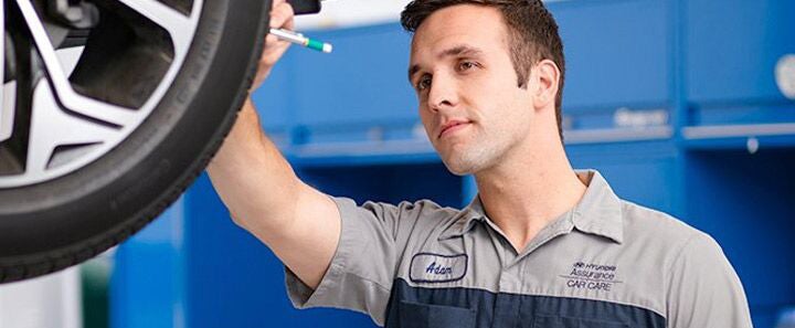 A mechanic in a automotive service uniform inspects a vehicle’s raised wheel, holding a tool up close to the tire. He appears focused while working in a brightly lit service bay with blue cabinets in the background. A name tag with text is visible on his shirt.