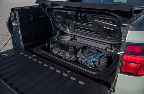 The open truck bed of a Hyundai Santa Cruz, showcasing its versatile cargo space. The bed features a lockable, weatherproof storage compartment under the main bed floor, with two backpacks visible inside.