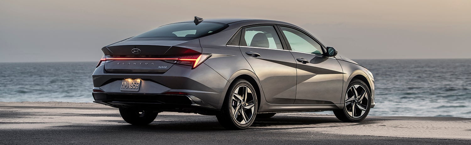 A gray Hyundai Elantra parked on a beach with the ocean in the background. The sun is setting, creating a warm glow.