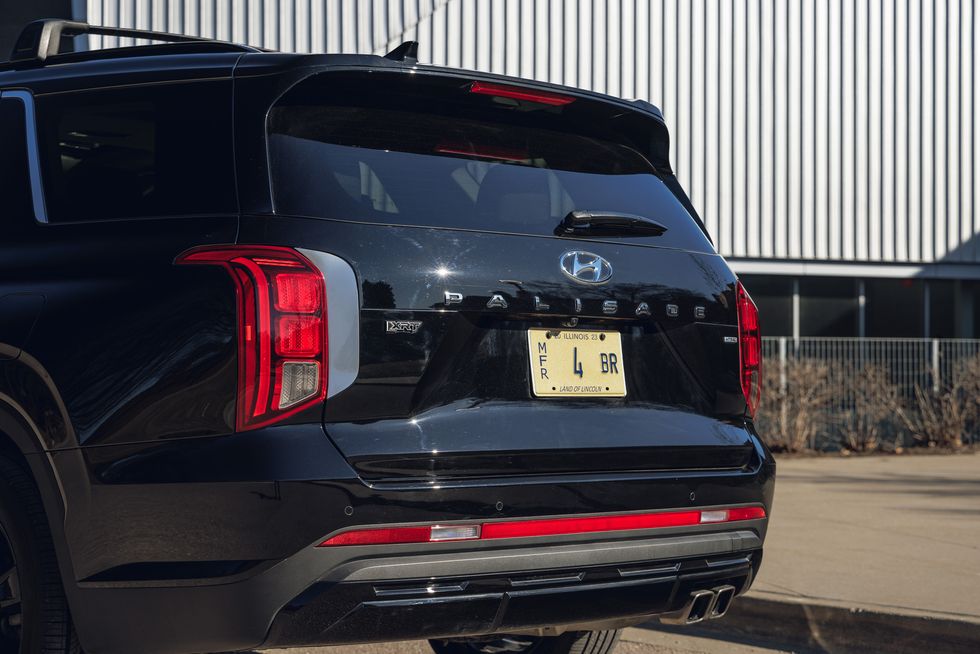 A rear view of a black Hyundai Palisade parked in front of a modern building. The image highlights the vehicle's distinctive taillights, "Palisade" badge, and dual exhaust tips.