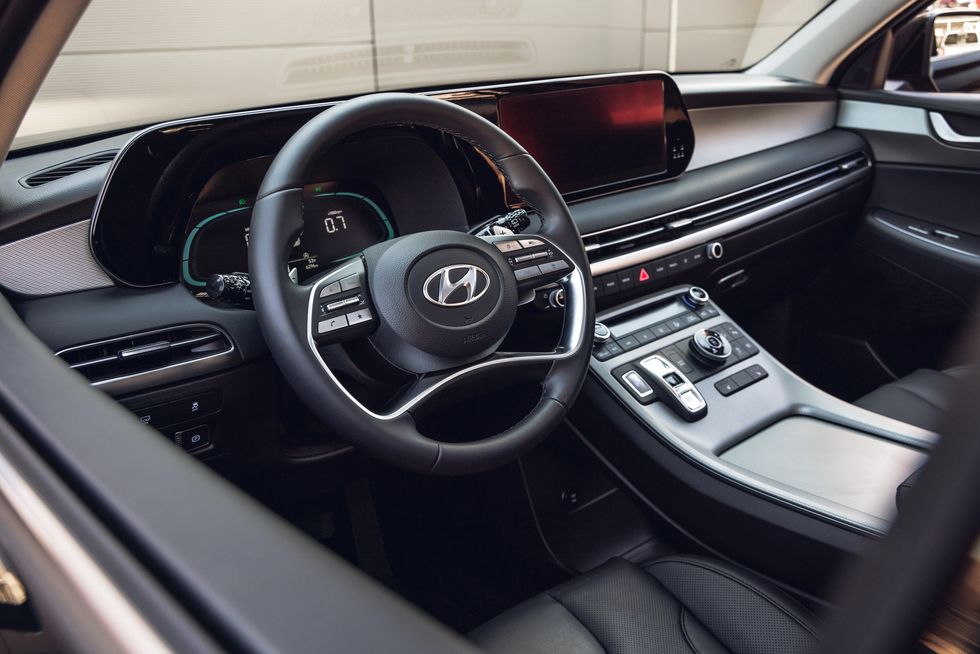 The interior of a Hyundai Palisade, showing the driver's seat, steering wheel, and dashboard with a large touchscreen display. The digital instrument cluster displays a charge level indicator.