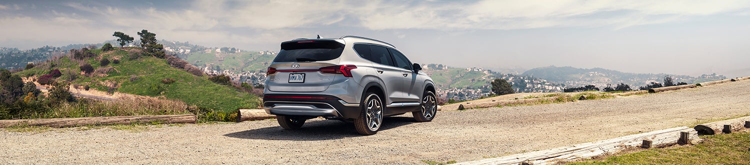 A silver Hyundai Santa Fe parked on a dirt road overlooking a scenic landscape with rolling hills and a city in the distance.