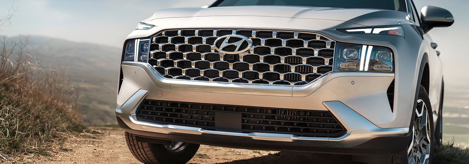 A close-up of the front of a silver Hyundai Santa Fe. The image highlights the vehicle's distinctive grille and headlights, showcasing its rugged and stylish design.