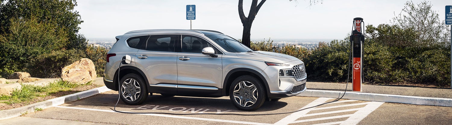 A silver Hyundai Santa Fe PHEV is parked at an EV charging station. The vehicle is plugged in and charging.