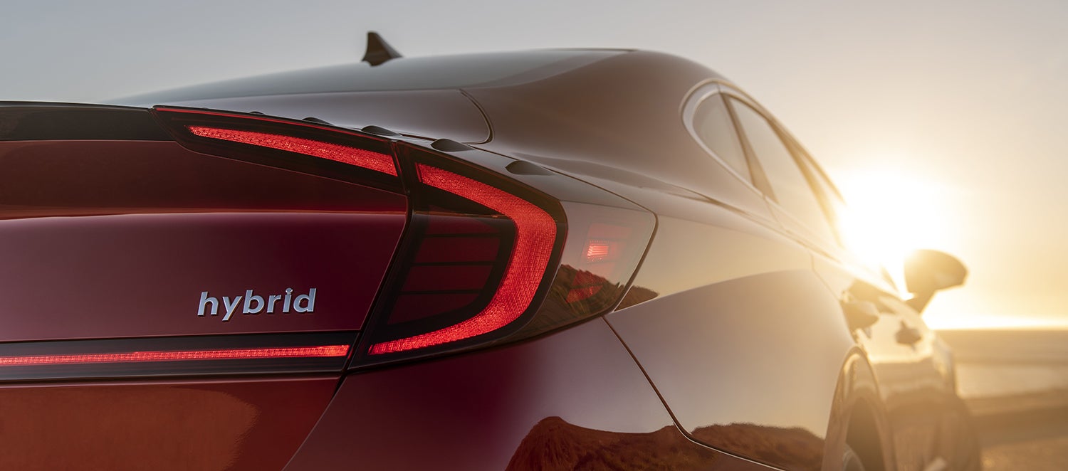 A close-up of the rear of a red Hyundai Sonata Hybrid at sunset. The image highlights the distinctive taillights and the "hybrid" badge.