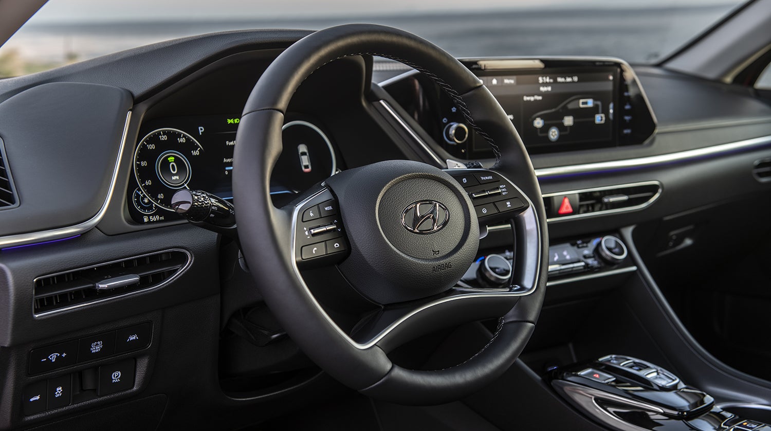 The interior of a Hyundai Sonata Hybrid, showing the driver's seat, steering wheel, and dashboard with a large touchscreen display. The digital instrument cluster displays a hybrid vehicle-specific gauge.