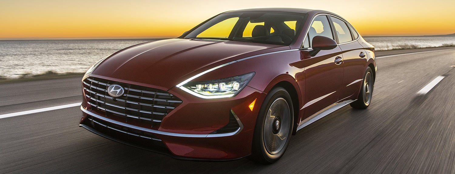 A red Hyundai Sonata driving along a coastal road at sunset, with the ocean visible in the background.
