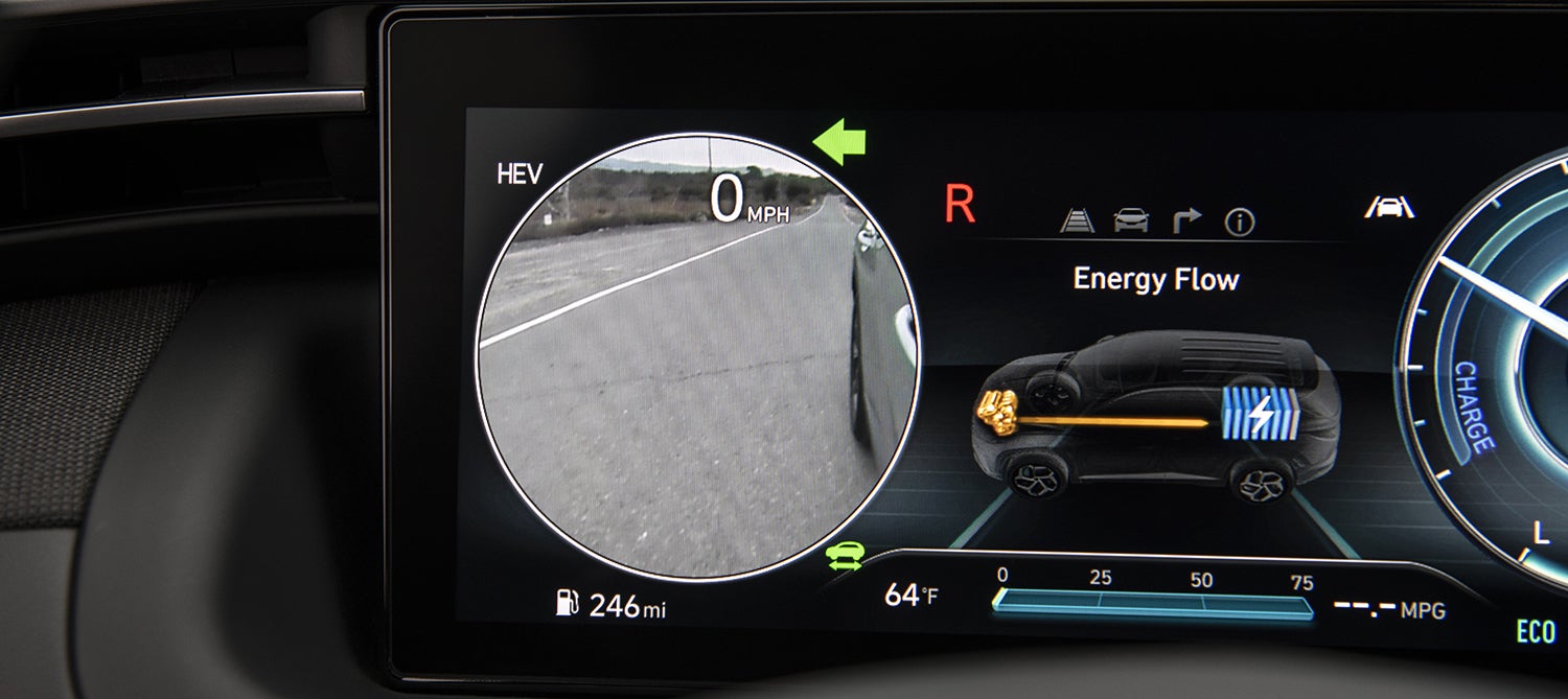 A close-up of the digital instrument cluster in a Hyundai Tucson Plug-in Hybrid. The display shows the current speed (0 mph), a forward-facing camera view, and an energy flow diagram indicating the vehicle is currently operating in hybrid mode.