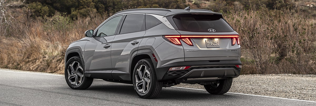 A rear three-quarter view of a silver Hyundai Tucson driving on a winding road with hills and trees in the background.