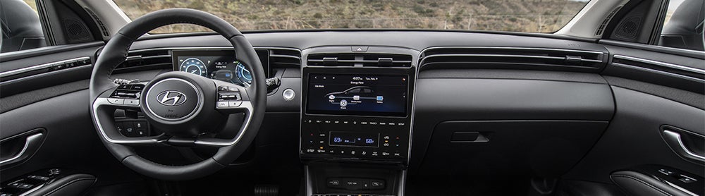 A close-up view of the interior of a Hyundai Tucson. The image shows the driver's seat, steering wheel, and dashboard with a large touchscreen display and a digital instrument cluster.